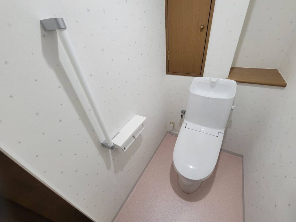 福岡市西区でトイレリフォームした施工後の画像。LIXILアメージュシャワートイレに交換しました。 これまで手すりがなかったた部分に下地工事を行い、新しく手すりを設置しています。床クッションフロアはピンクでの花柄のデザイン。 壁クロス星柄。天井にもおすすのデザイン。