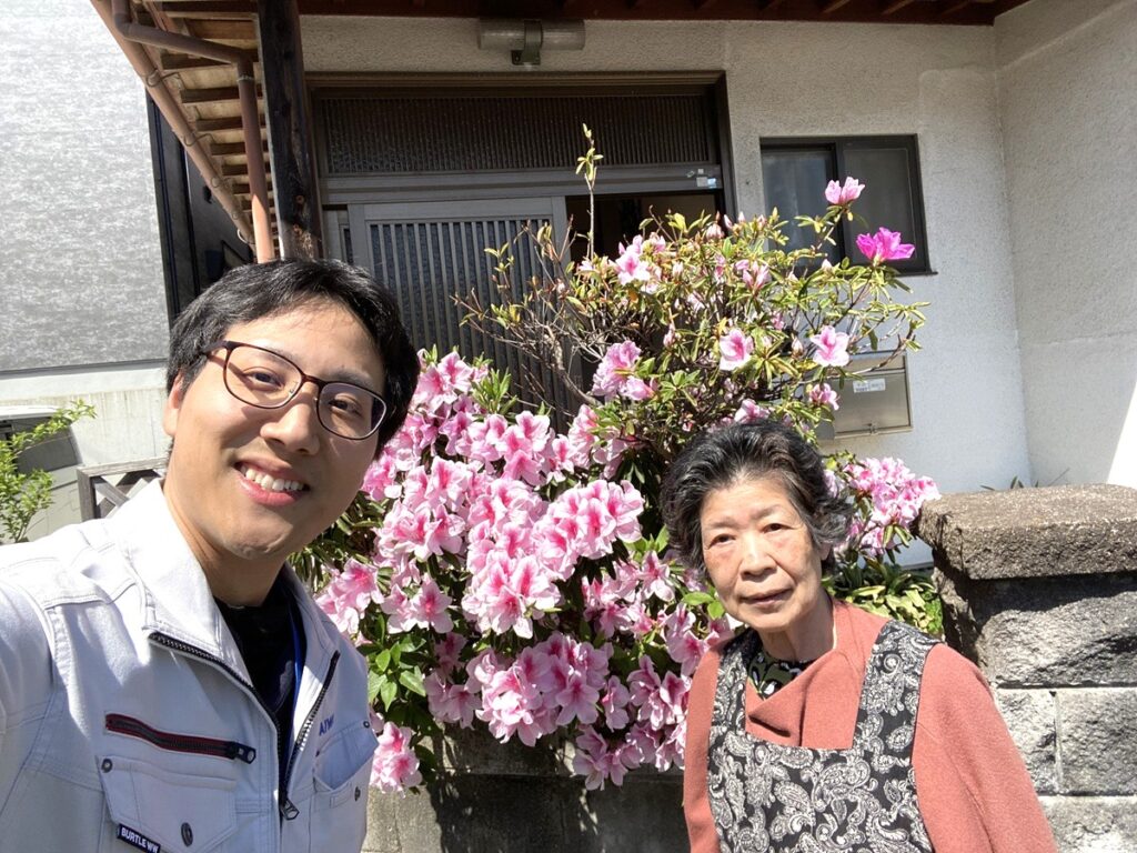 福岡市南区でガス給湯器交換のご依頼をいただいたお客様とつつじの前で撮影している画像。