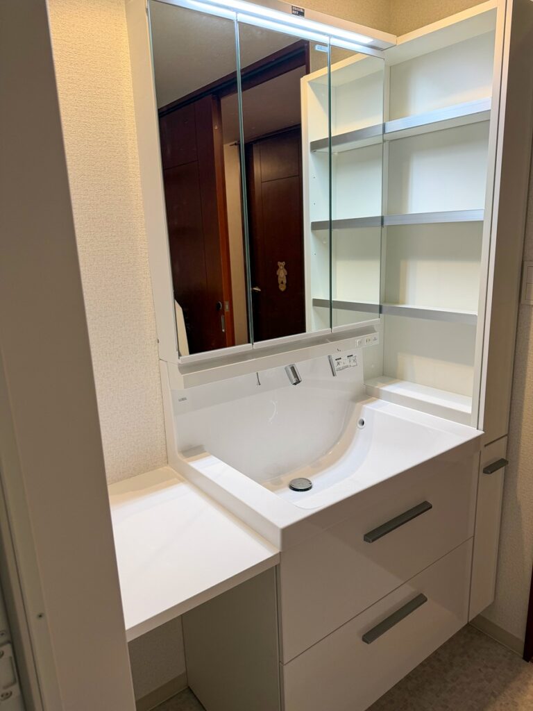 福岡市南区で洗面化粧台をリフォームした施工後の画像。LIXILEVの大和オリジナル洗面化粧台。色は白。洗面化粧台の隣には15㎝幅の同シリーズのトールタイプの収納棚を設置。三面鏡裏とトール収納で収納力がアップしました。アイロンをかけれる台も設置して便利そうな洗面室です。