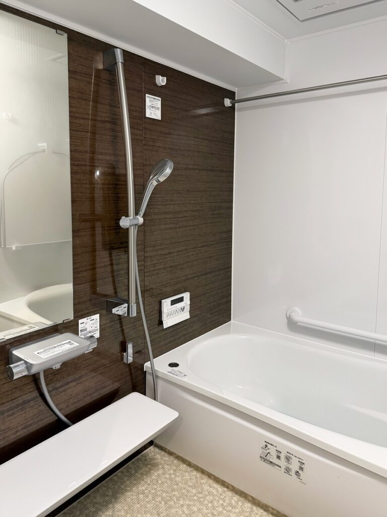 福岡市中央区で浴室リフォームをした施工後の画像。ダークブラウンの木目調のアクセントパネルの高級感と床のラグ調のベージュの床がおしゃれです。